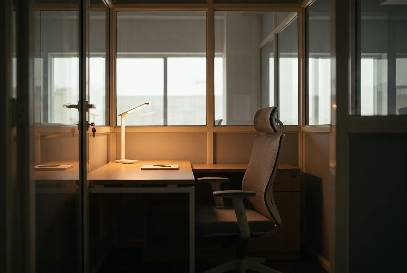 A single empty desk in a dim office bay with one lamp left on